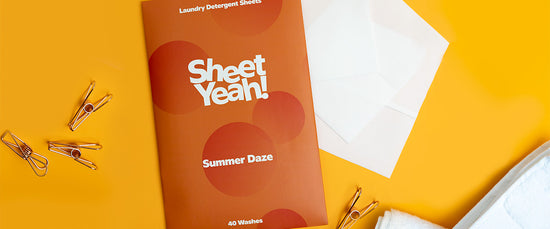 A single pack of Summer Daze Sheet Yeah! Laundry Detergent Sheets are laying on an orange background with metal pegs, white laundry sheets and a white fluffy towel spread around it.