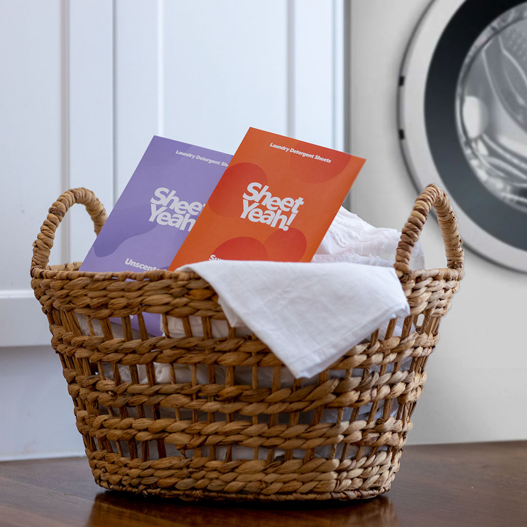 A laundry basket filled with fluffy clean towels sitting on the floor in front of a washing machine. Two packs of Sheet Yeah! laundry detergent sheets are on top of the towels, showing an alternative eco-friendly option for doing laundry.