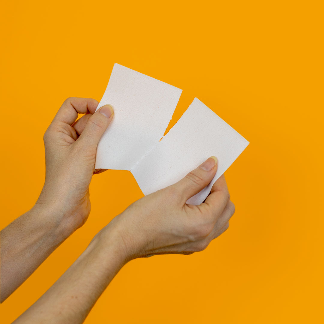 A pair of hands tearing a Sheet Yeah! Laundry Detergent Sheet in half along the perforated line on yellow background
